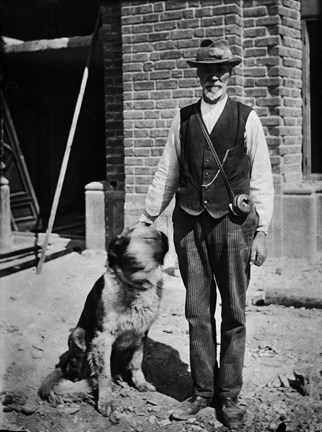 Svart-vit bild med en stående man och en sittande hund. Mannens ansikte är delvis skuggat, han bär skjorta, hatt, smårandiga byxor och skor. Hundens ansikte är suddigt i bilden eftersom han rör sig. I bakgrunden syns en murad vägg. 