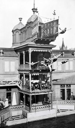 Ett högt hopptorn i trä vid en bassäng. Flera personer hoppar från avsatserna. Nedanför tittar publiken på.