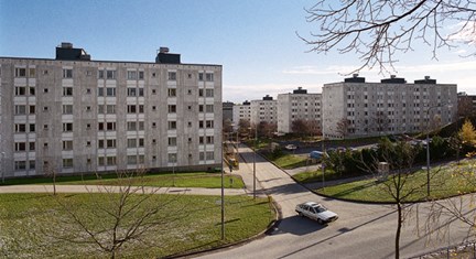 Flervåningshus på rad och öppna gräsplaner i förgrunden. En bil svänger ut på en väg.