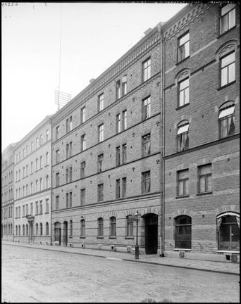 Svart-vit bild av en husfasad i tegel med fem våningar. Högst upp på taket syns en antenn.