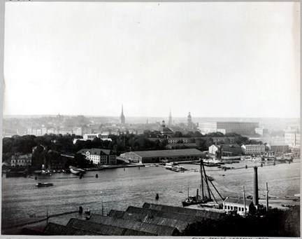 En utsiktsbild mot Skeppsholmen. Här syns ett gammalt badhus, förråd och båthus. I bakgrunden Gamla stan och slottet.  