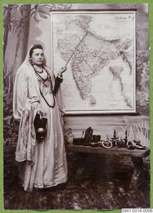 En kvinna i indisk dräkt står framför en karta över Indien och pekar med en pekpinne. Intill henne finns en låg bänk med olika konsthantverksprodukter.