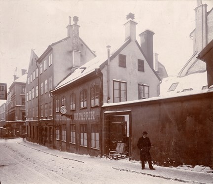 En snöig gata. En man står på trottoaren, en annan är på väg ut från gården genom en port, med ett paket på en sparkstötting. På fasaden: Linderoths urfabrik. Mellan orden sitter en klocka.